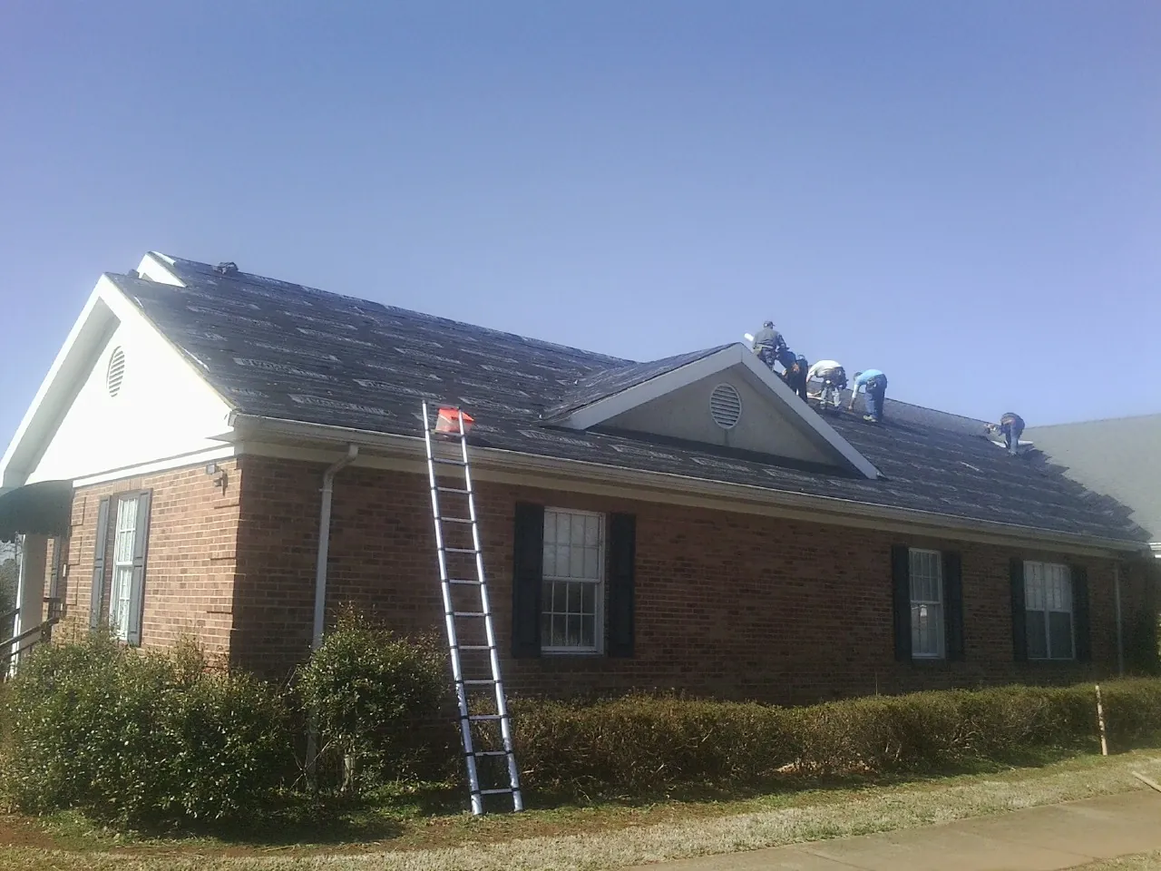 Crew tearing shingles off a brick ranch roof with a ladder set — Metal Roof Repair work by our crew in Arrington