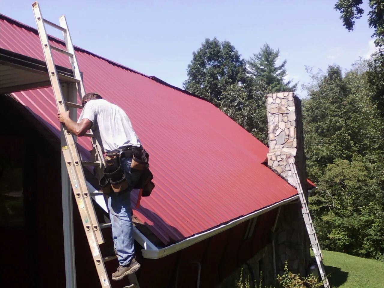 Roofer on a ladder beside a red metal roof and stone chimney — Soffit & Fascia Repair by Roofing Brothers in Arrington